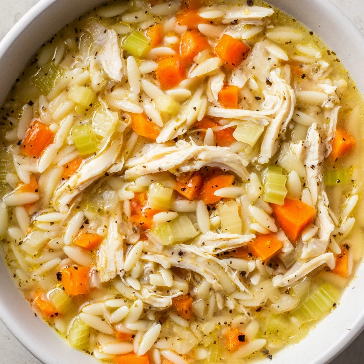 A close-up of Lemon Pepper Chicken Orzo Soup in a rustic bowl, garnished with fresh parsley and black pepper, alongside crusty bread for dipping.  