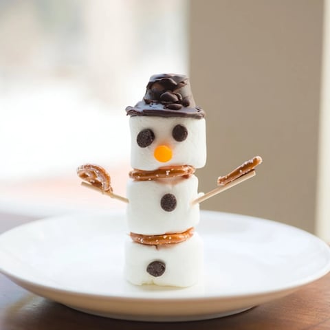 Marshmallow snowman stick arrangement with smiling faces, a fun winter dessert with pretzels for arms.