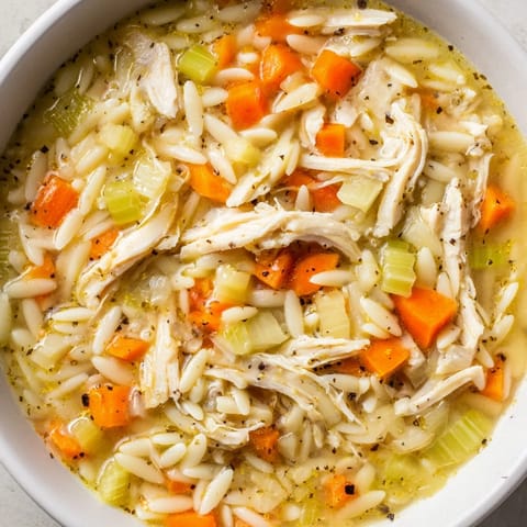 A close-up of Lemon Pepper Chicken Orzo Soup in a rustic bowl, garnished with fresh parsley and black pepper, alongside crusty bread for dipping.  